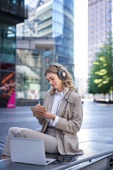 Vertical shot of smiling adult woman studying outdoors, listening to online course, taking notes, attend team meeting via app in laptop, using headphones.