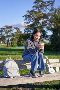 Young woman sitting in park on bench with ukulele, looking at smartphone, reading message on mobile phone and smiling.