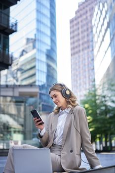Corporate woman in suit, sitting in city, listening to music with laptop, scrolling news on her mobile phone, relaxing on lunch break.