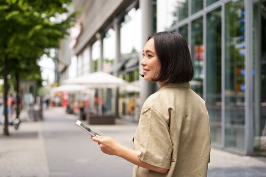 Rear shot of young woman walking in city, going down the street and smiling, holding smartphone. View from behind.
