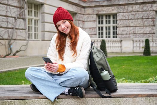Redhead girl, female student sits with mobile phone on bench in parj, leans on her backpack. Woman browsing social media app feed on her smartphone.