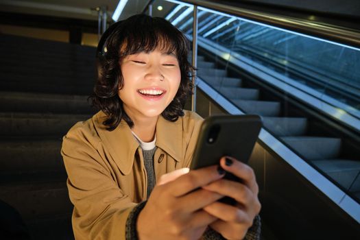 Close up portrait of smiling asian girl student, listens music in headphones and looks at mobile phone, uses smartphone, sits on staircase in public place.