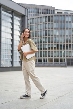 Vertical portrait of stylish asian girl student, talking on mobile phone while walking, holding laptop.