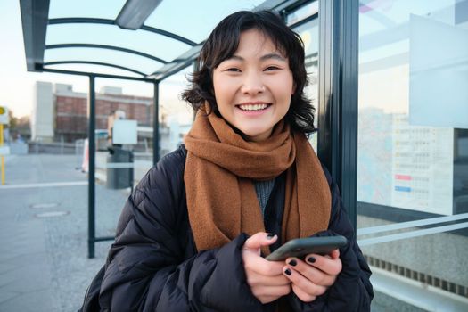 Beautiful young woman, standing on bus stop, waiting for her public transport, using mobile phone, using smartphone application.