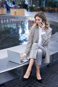 Vertical shot of corporate woman sitting outdoors with laptop, talking on mobile phone, working ouside office building while waiting for someone.
