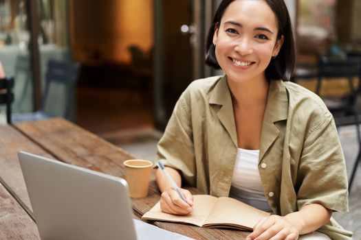 Portrait of young korean girl making notes, listening online meeting, lecture, looking at laptop screen, working remotely from cafe.