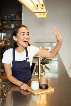 Smiling asian girl barista calling client, giving out order in cafe, prepared batch brew filter coffee, wearing apron behind counter.