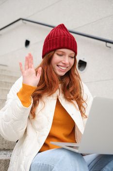 Portrait of happy young woman, redhead girl with laptop, waves hand and says hello on video chat, has conversation on computer application, talks to someone.