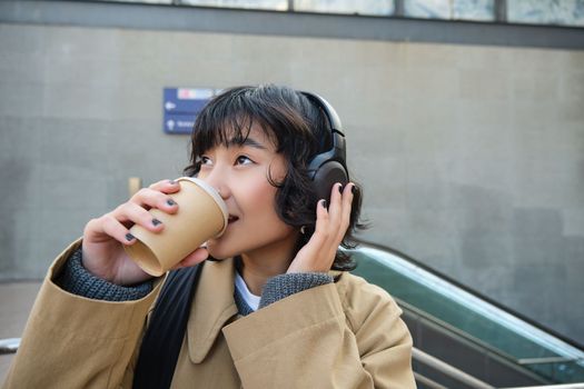 Portrait of stylish asian woman in headphones, drinks coffee to go and smiles, enjoys cappuccino while commutes, stands on street.