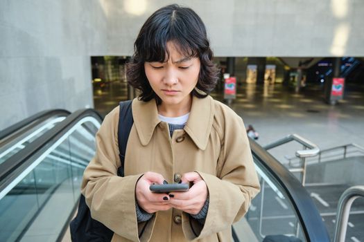 People in city. Portrait of girl looks concerned at smartphone screen with frowned worried face expression. Woman goes up escalator with mobile phone.