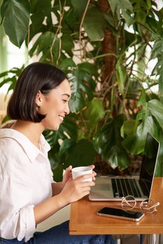 Vertical shot of beautiful woman sits in cafe with laptop, works or studies online at co-working space, smiling relaxed.