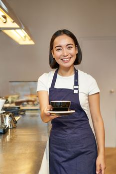 Smiling beautiful asian girl barista, working in cafe, holding cup of tea, giving client their order, wearing uniform apron.