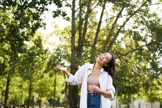 Happy young asian woman walking alone, dancing and singing in park, smiling carefree. People and lifestyle concept.