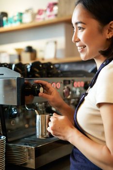 Happy asian woman, barista using coffee machine to make order, steaming milk for cappuccino and latte, laughing and smiling while working in cafe.