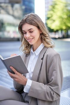 Smiling woman in suit writing down ideas, using notebook and pen, sitting outdoors in city centre, working.