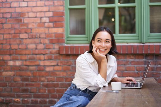 Portrait of young stylish woman, influencer sitting in cafe with cup of coffee and laptop, smiling and looking confident.