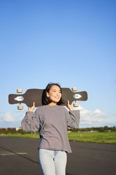 Vertical shot of carefree asian girl with longboard. Young woman skater holding cruiser on her shoulders and walking on road, skateboarding.