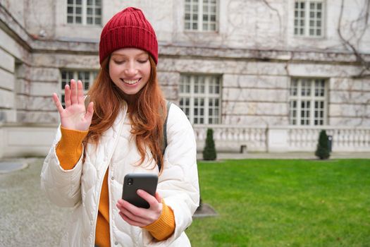Portrait of smiling redhead woman says hello, video chats with friend on smartphone app, waves hi at mobile camera, stands on street.
