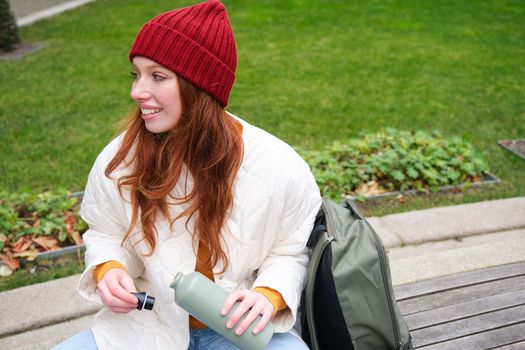 Portrait of young redhead woman in hat, sitting on bench, holding thermos, drinking hot drink from flask and smiling, resting in park.