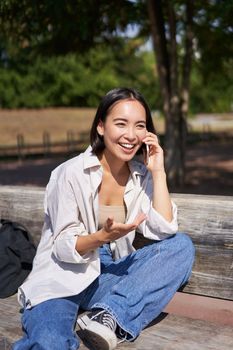 Portrait of happy young asian woman talking on mobile phone in park, sitting on bench and having a telephone call, chatting lively.