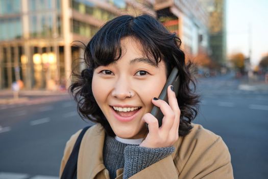 Portrait of happy korean girl, talks on mobile phone, looks surprised and happy, receives positive great news over telephone conversation, stands on street of city.