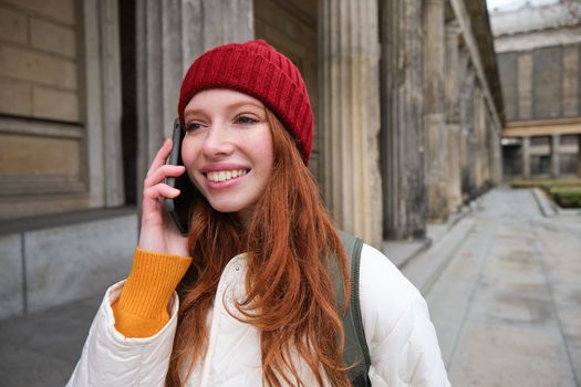 Smiling redhead female tourist talks on mobile phone and walks around city. Happy student in red hat calls friend, stands on street and uses smartphone.