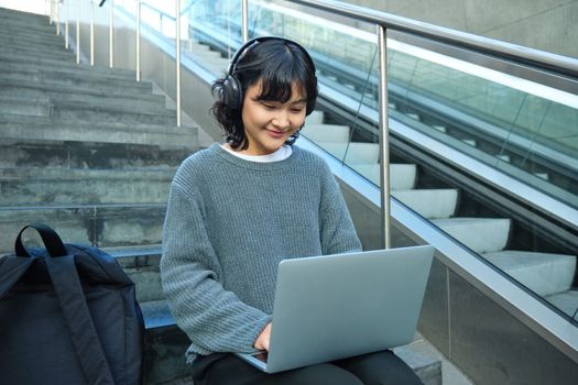 Portrait of beautiful young korean woman, student sits on stairs in public place, listens music in headphones, works on project on laptop, works remotely.