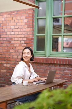 Portrait of stylish young woman, brunette girl with laptop, sitting outdoors and using computer.