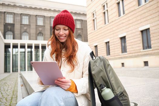 Redhead girl smiles, sits outdoors near building with digital tablet, thermos and backpack, connects to public internet and searches smth online on her gadget.