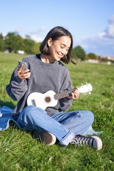 Vertical shot of smiling korean girl sitting in park with ukulele and smartphone, using mobile app to tune instrument, looking for chords while playing.
