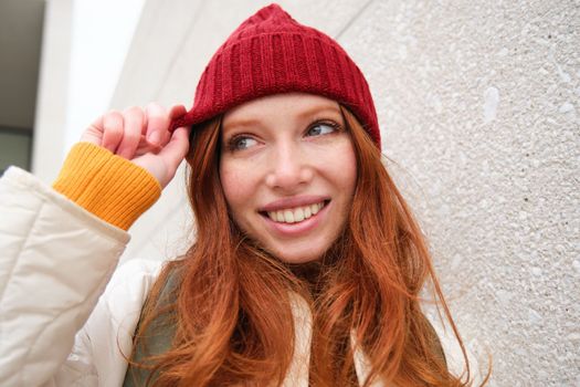 Stylish redhead girl in red hat, smiles and looks happy, poses outdoors on street, looks relaxed and lively.