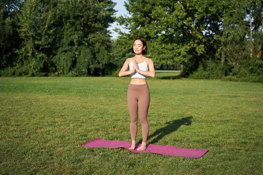 Mindfulness and wellbeing. Young woman doing yoga, standing on mat in park, making asana, meditating on fresh air, urban life concept.