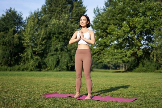 Young asian woman in sportswear, holding hands in namaste gesture, practice yoga, meditating and relaxing on fresh air in park, concept of mindfulness and wellbeing.