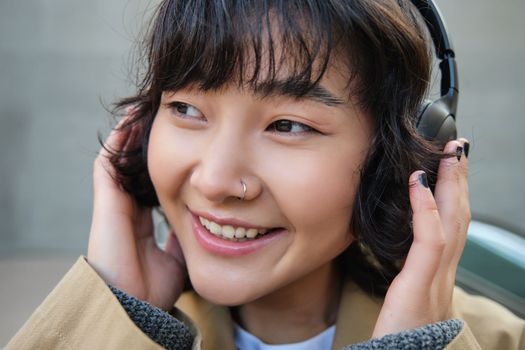 Beautiful smiling korean girl in headphones, wondering around town, standing on street and smiling, listening to music.