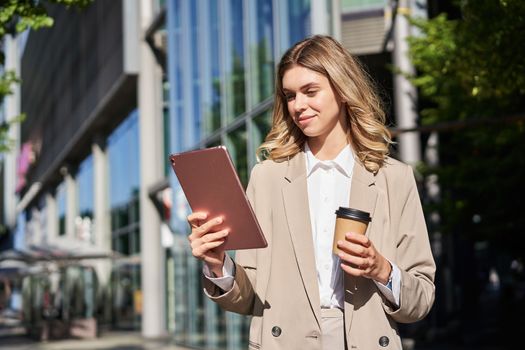 Portrait of young confident businesswoman on street, drinks her coffee and looks at tablet, works on her way to office. Digital nomad concept.