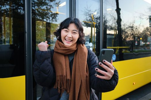 Enthusiastic asian woman, standing on bus stop with smartphone, looking at phone screen with amazed, triumphing face, winning, hear great news on video chat.