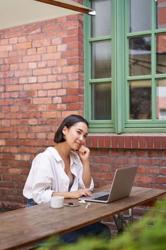 Beautiful brunette woman sitting outdoors and using laptop, chatting online, working with computer outside office.