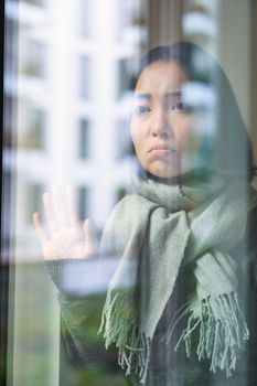 Asian woman feels unwell, stays at home, looks outside window with jealous, distressed face, wants go outside but caught cold.