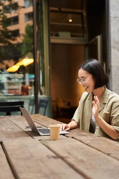 Portrait of asian woman looking at laptop, video chat, talking with someone via computer camera, sitting in cafe and drinking coffee, online meeting.
