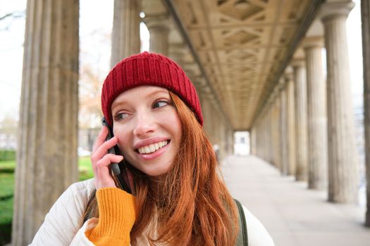 Smiling cute redhead woman makes a phone call, holds telephone near year, has mobile conversation, using smartphone on street.