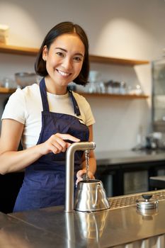Vertical shot of barista, girl in coffee shop, pouring water in kettle, preparing order in cafe, smiling at camera.