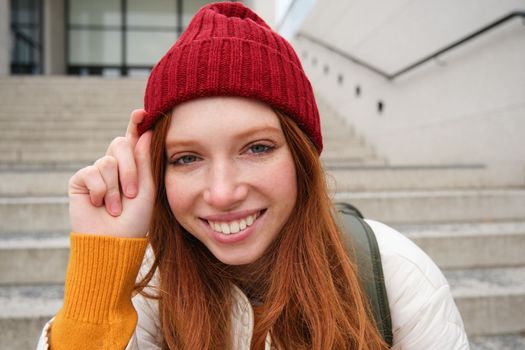 Stylish redhead girl in warm red hat, smiling relaxed, sitting with backpack on stairs near building, waits for someone outdoors.