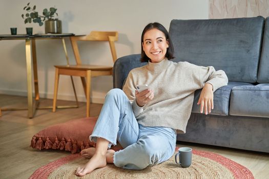 Portrait of stylish korean woman sits on floor with smartphone, using mobile phone, smiling pleased, concept of staying at home and relaxation.