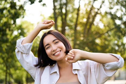 People lifestyle. Portrait of young brunette woman dancing, smiling and laughing, walking in park with hands lift up high, enjoying summer day outside.