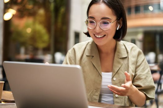 Online meeting. Young urban girl, asian woman talking with laptop, video chat, gesturing, sitting in an outdoor cafe and working remotely.