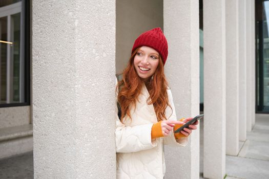 Stylish urban girl using mobile phone app, standing in city, waiting for taxi, looking at smartphone application, texting message.