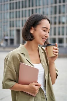 Vertical shot of beautiful korean girl drinks coffee, holds tablet, poses on street of city. Copy space