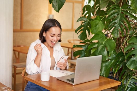 Joyful young asian woman winning, celebrating victory or achievement, receive good news on mobile phone, sitting with laptop and working.