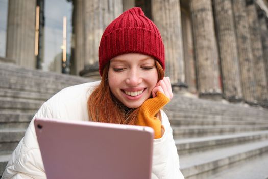 Portrait of young redhead woman sitting outdoors on stairs, reading e-book on digital tablet, wearing red hat and warm clothes.