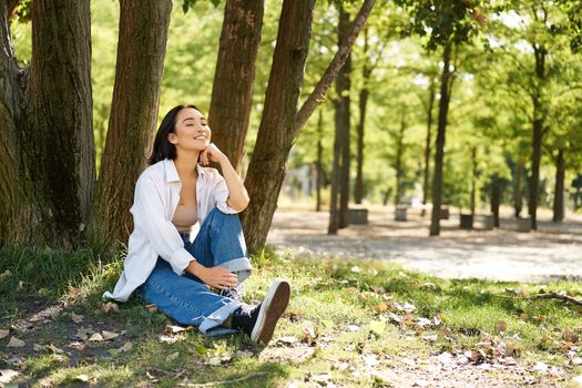 Relaxed young woman, resting near tree, sitting in park on lawn under shade, smiling and looking happy, walking outdoors on fresh air.
